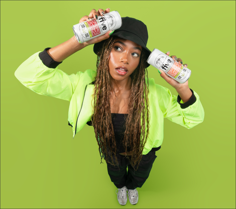 Young woman in neon green jacket and black bucket hat holds two 'five' beverage cans above her head against lime background.