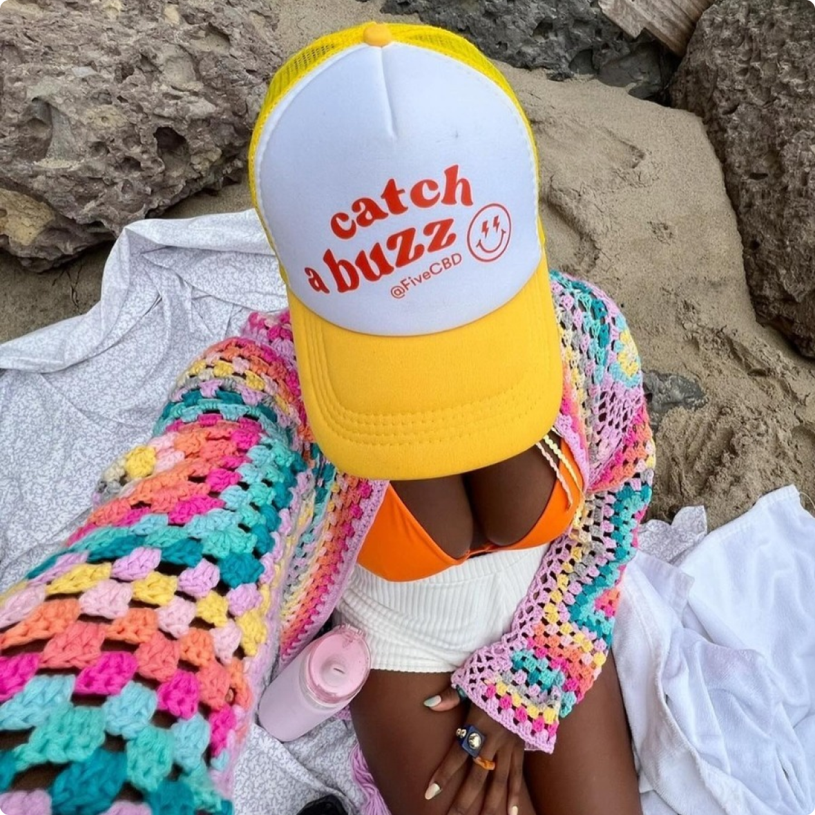 Top-down selfie of person wearing yellow trucker hat reading 'catch a buzz @FiveCBD', colorful crochet and orange bikini