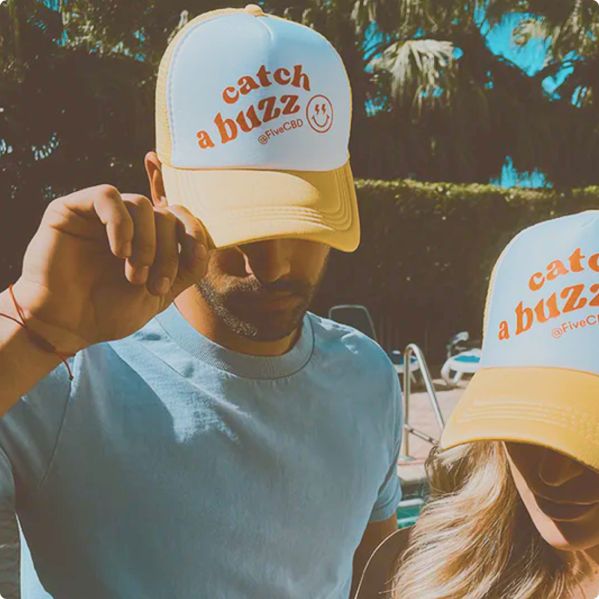 Two people wearing yellow-and-white trucker hats embroidered catch a buzz @FiveCBD, hats partially shading their faces