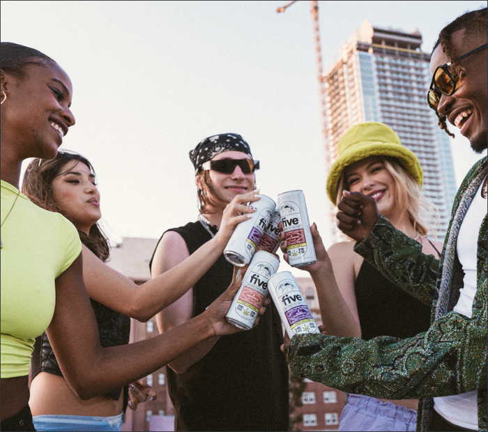 Five young adults toasting with cans labeled 'five' in an urban outdoor setting, smiling and enjoying themselves.