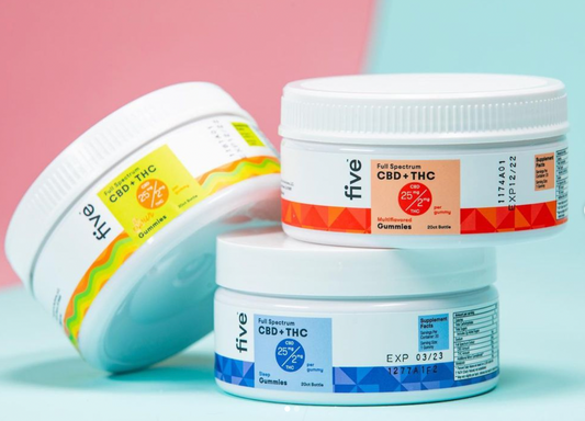 Three stacked tubs of Five full‑spectrum CBD and THC gummies showing product labels and dosage, on a pastel pink and teal background