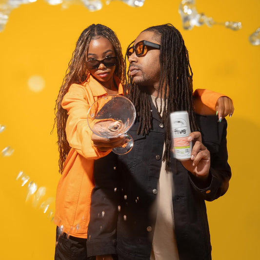 Two people in sunglasses pose against a yellow background, woman holds a splashing glass and man holds a Five THC seltzer can